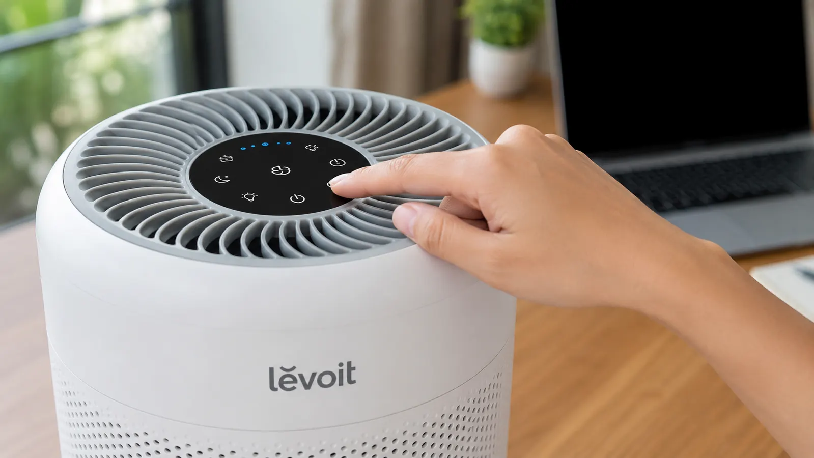 A hand pressing the sleep mode button on the top panel of the Levoit air purifier next to a desk
