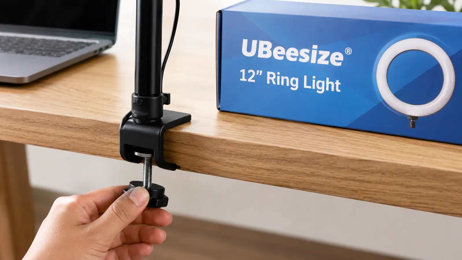 A hand adjusting the clamp of a UBeesize ring light onto the edge of a desk beside a laptop