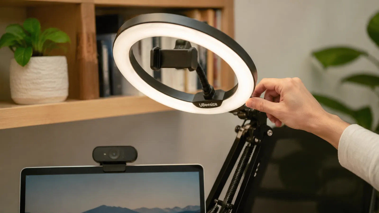 Hand adjusting the UBeesize ring light's angle on its clamp arm next to a laptop webcam